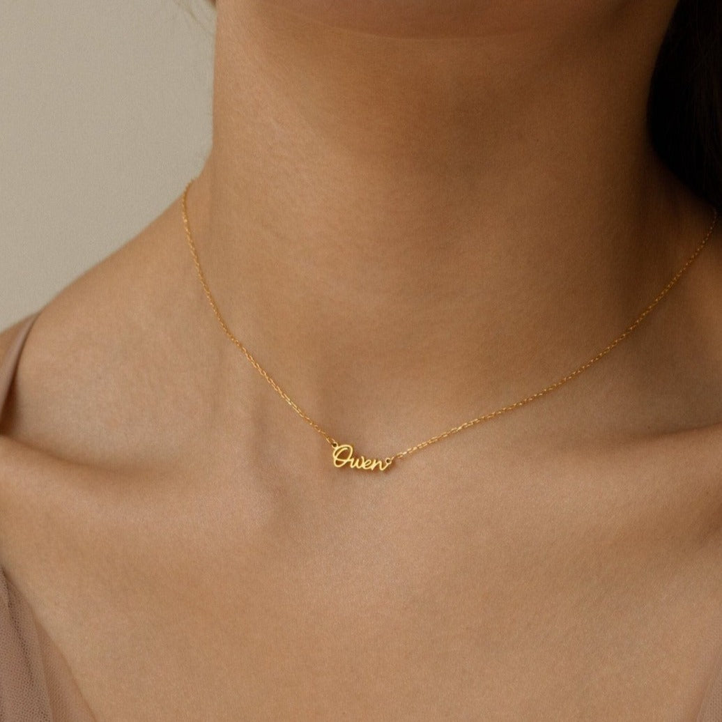 A woman wears the Tiny Grace Name Necklace, a gold necklace featuring a custom nameplate pendant.