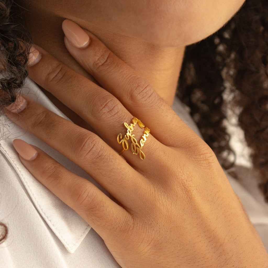 A hand with nude nails wears the Grace Duo Name Ring, a personalized gold ring in graceful cursive script, lightly resting on the collar of a white shirt.