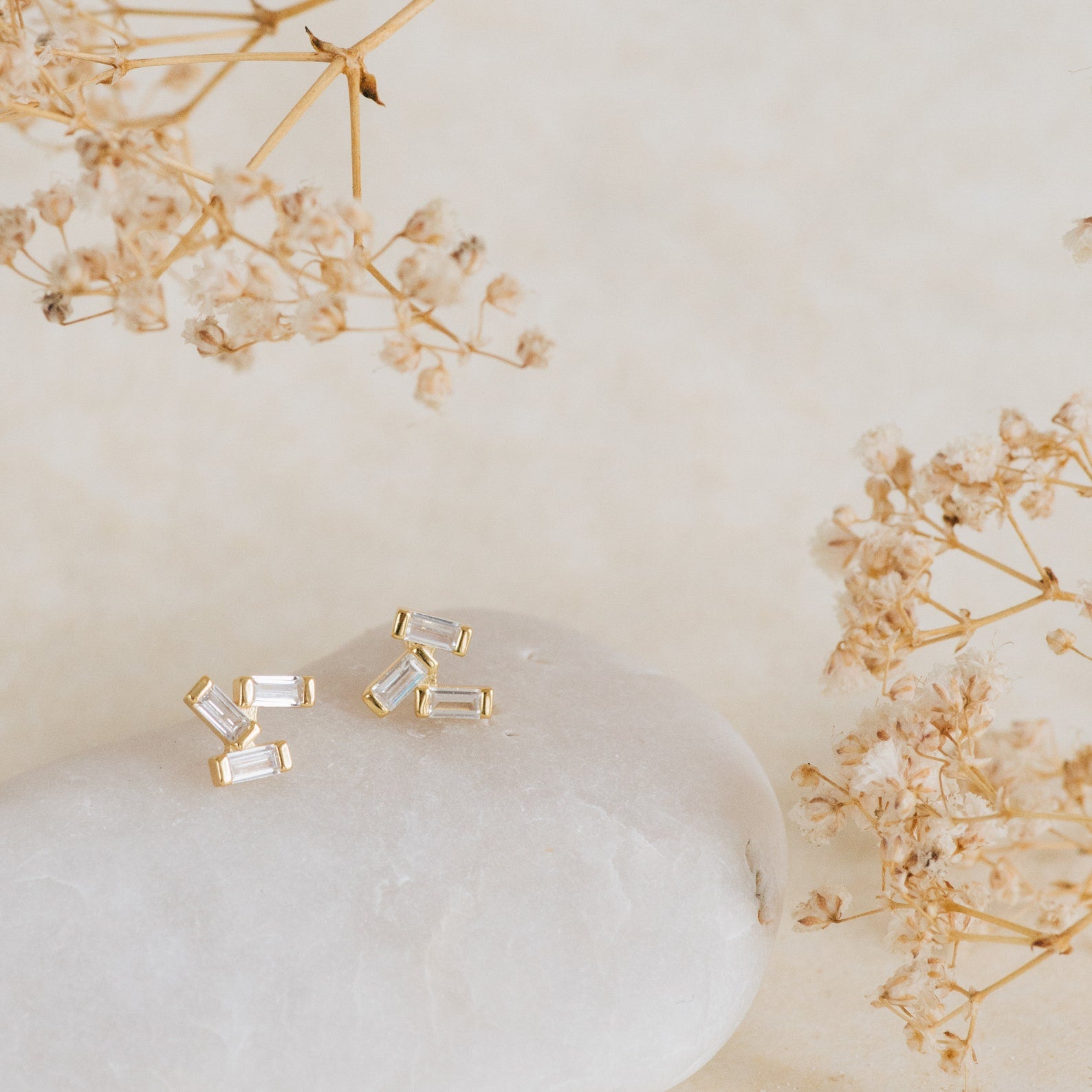 Glacier Diamond Studs sparkle atop a white stone, surrounded by dried beige flowers.