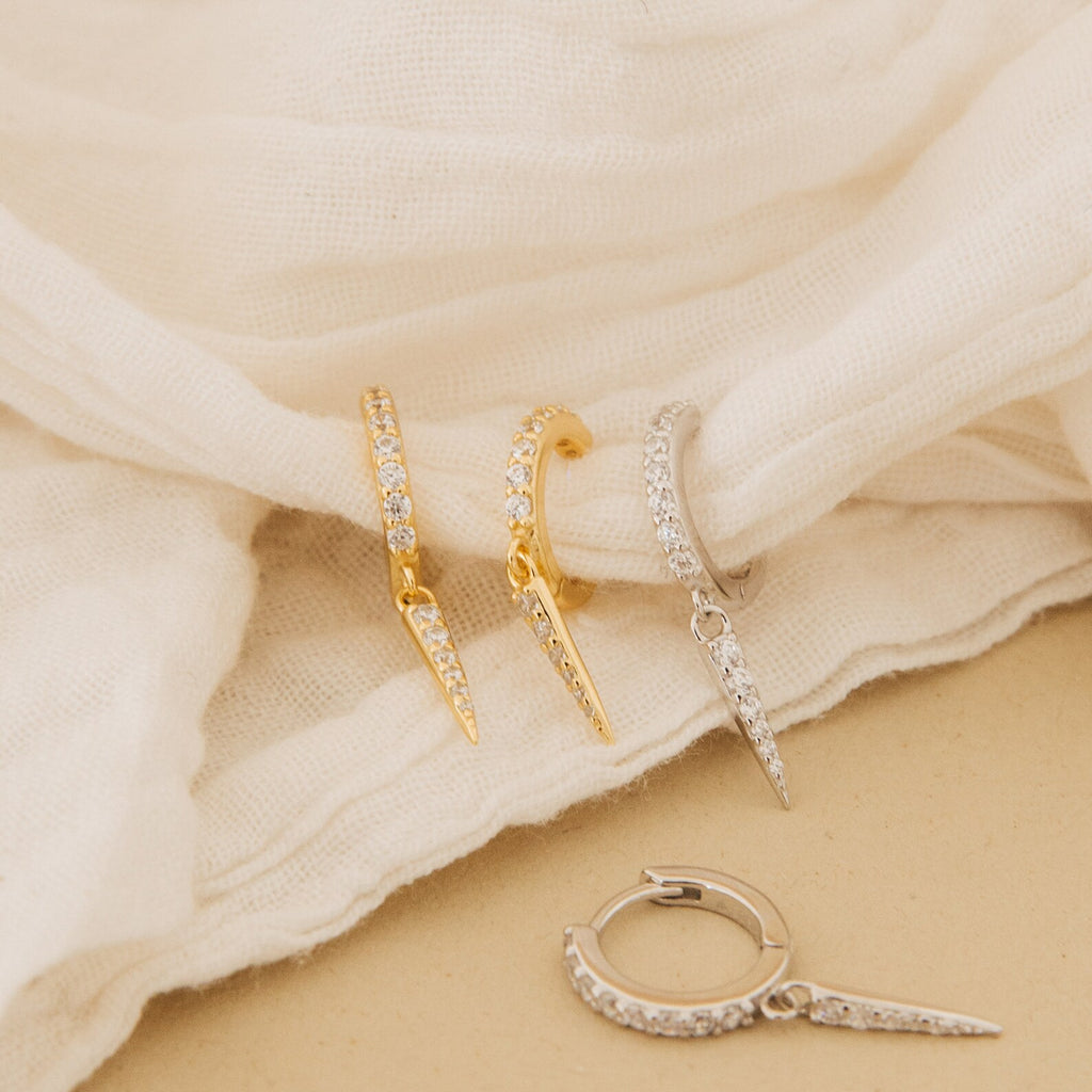 Quinn Pave Dagger Huggies—gold and silver edgy hoop earrings with crystals, inspired by pave dagger designs—displayed on soft cream-colored fabric.