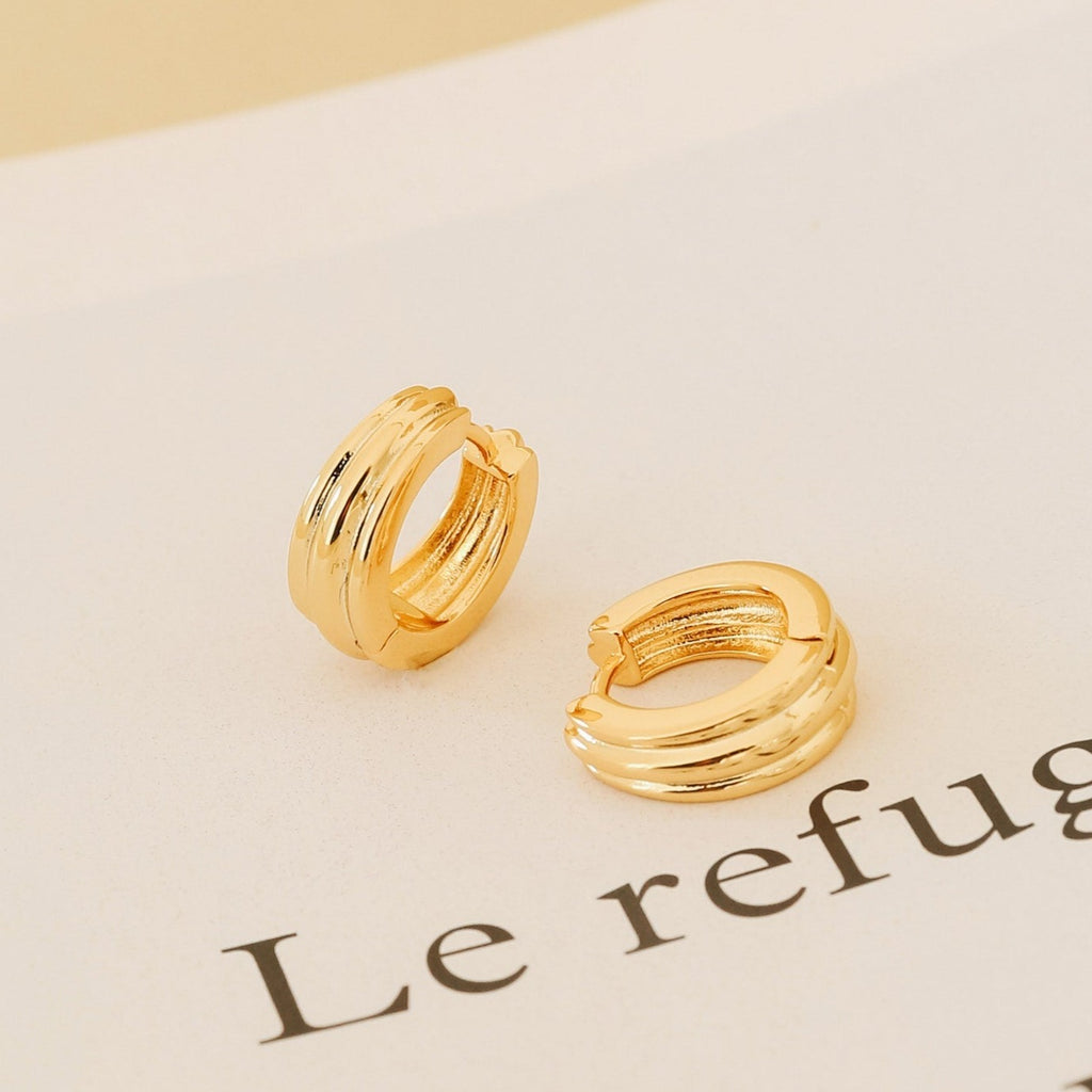 The Canal Chunky Hoops feature a layered gold design, resting on paper with partial text reading "Le refugi"—perfect as contemporary huggies or stylish everyday earrings.