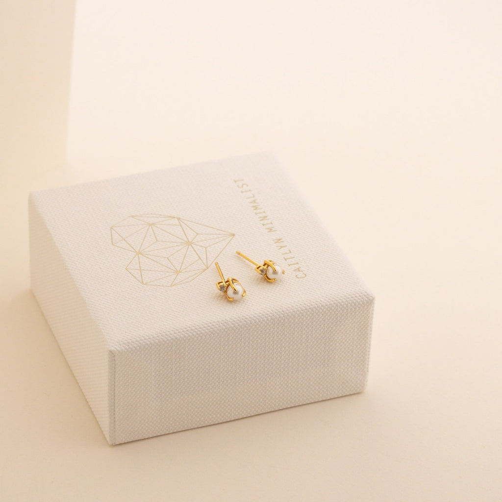 Small gold knot stud earrings with a geometric heart design rest on a white jewelry box, perfect for pairing with your Julia Pearl Studs.