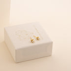 Small gold knot stud earrings with a geometric heart design rest on a white jewelry box, perfect for pairing with your Julia Pearl Studs.