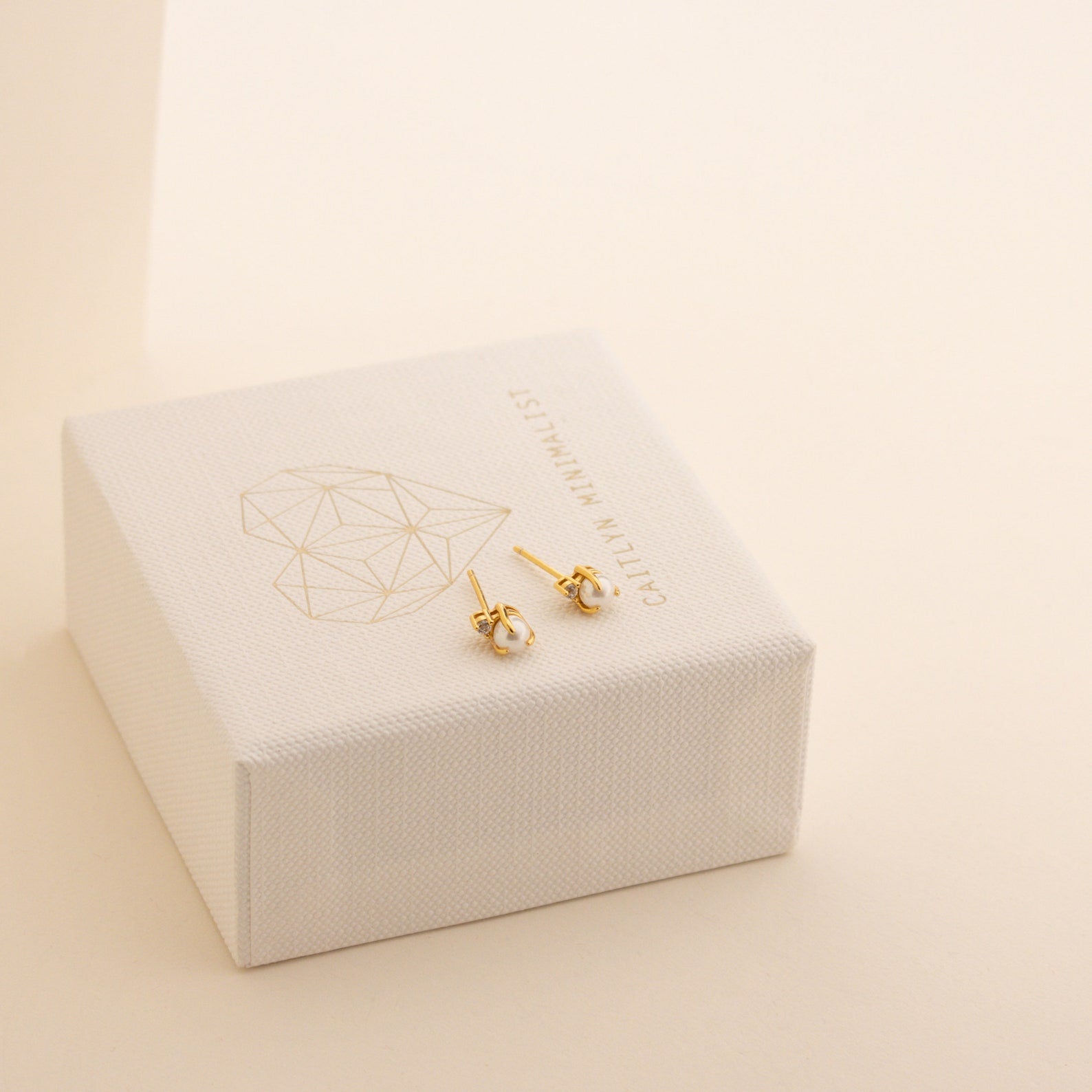 Small gold knot stud earrings with a geometric heart design rest on a white jewelry box, perfect for pairing with your Julia Pearl Studs.