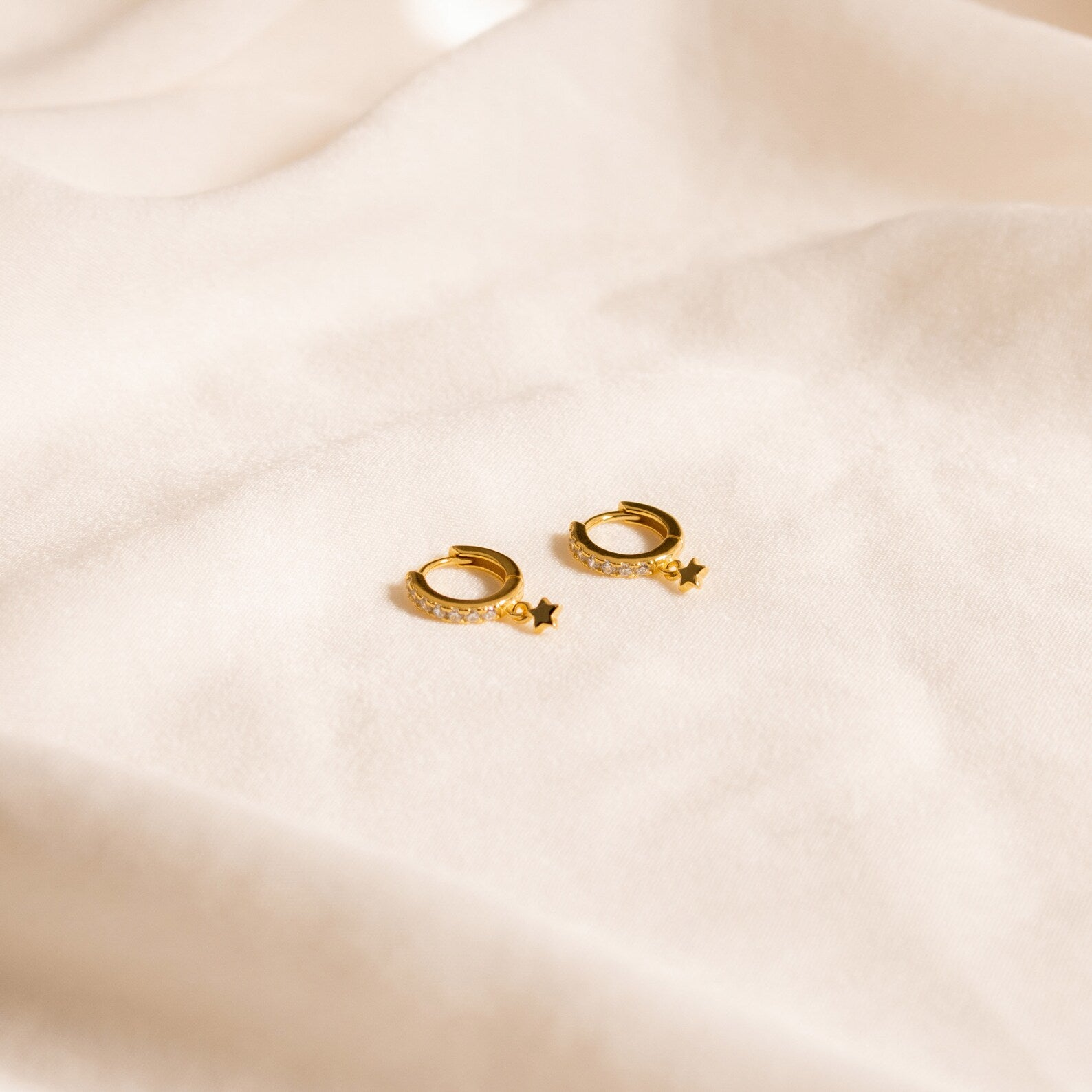 The Emily Pave Star Huggies, featuring crystal accents, are displayed on a soft, cream-colored fabric background, bringing a celestial touch to any look.