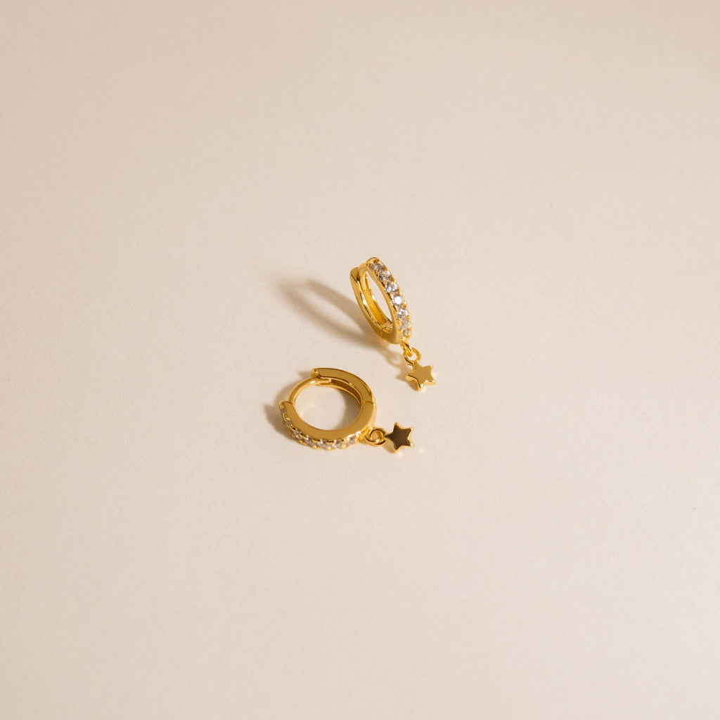 The Emily Pave Star Huggies feature gold hoops with star charms and embedded crystals, creating a celestial sparkle against a light beige background.