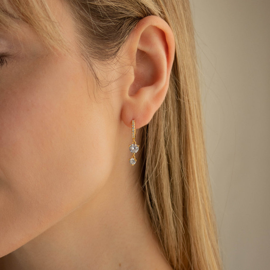 Close-up of a woman's ear wearing Caroline Pave Drop Huggies, gold embellished hoops featuring two dangling clear gemstones.
