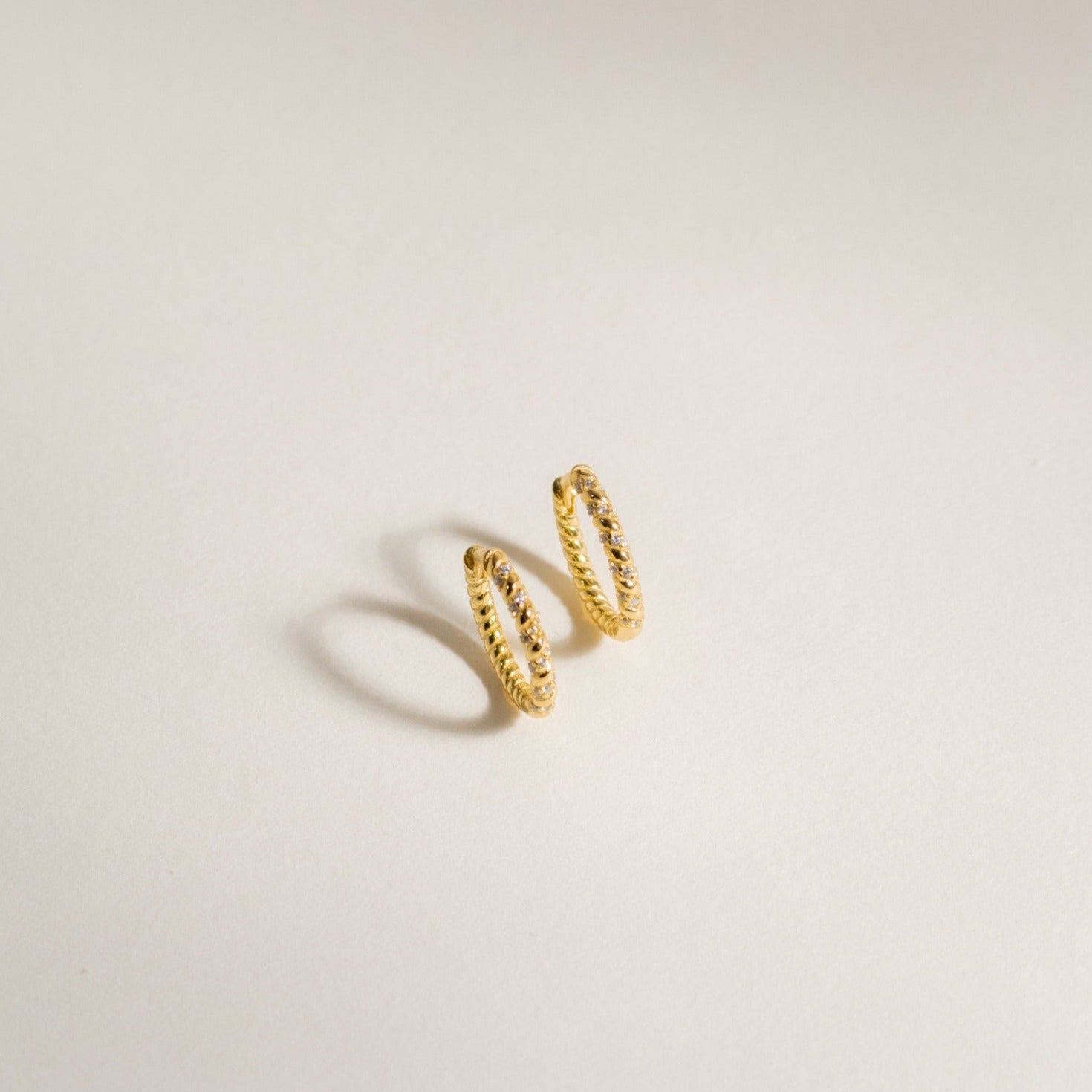 The Aster Pave Twisted Hoops are small gold hoops with a textured, twisted design that add subtle charm to any look. These dainty earrings are showcased on a simple, light-colored background.