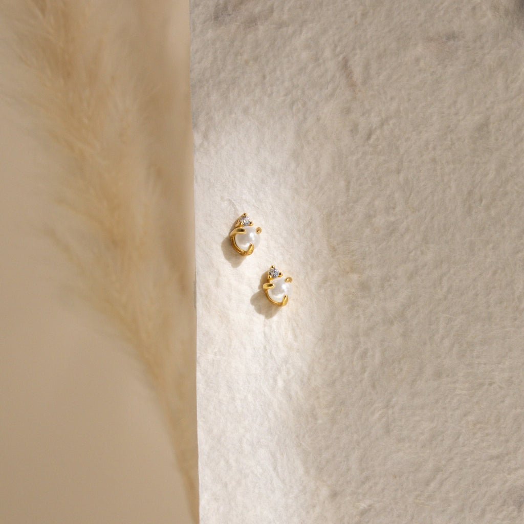 The Julia Pearl Studs—small gold earrings featuring crystals and freshwater pearls—are displayed on a textured cream background, softly lit from the left.