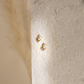 The Julia Pearl Studs—small gold earrings featuring crystals and freshwater pearls—are displayed on a textured cream background, softly lit from the left.