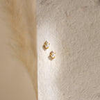The Julia Pearl Studs—small gold earrings featuring crystals and freshwater pearls—are displayed on a textured cream background, softly lit from the left.