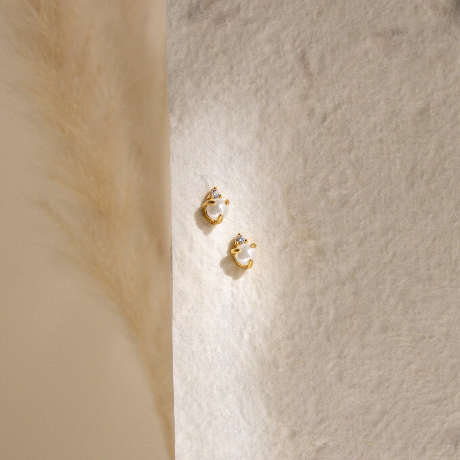 The Julia Pearl Studs—small gold earrings featuring crystals and freshwater pearls—are displayed on a textured cream background, softly lit from the left.