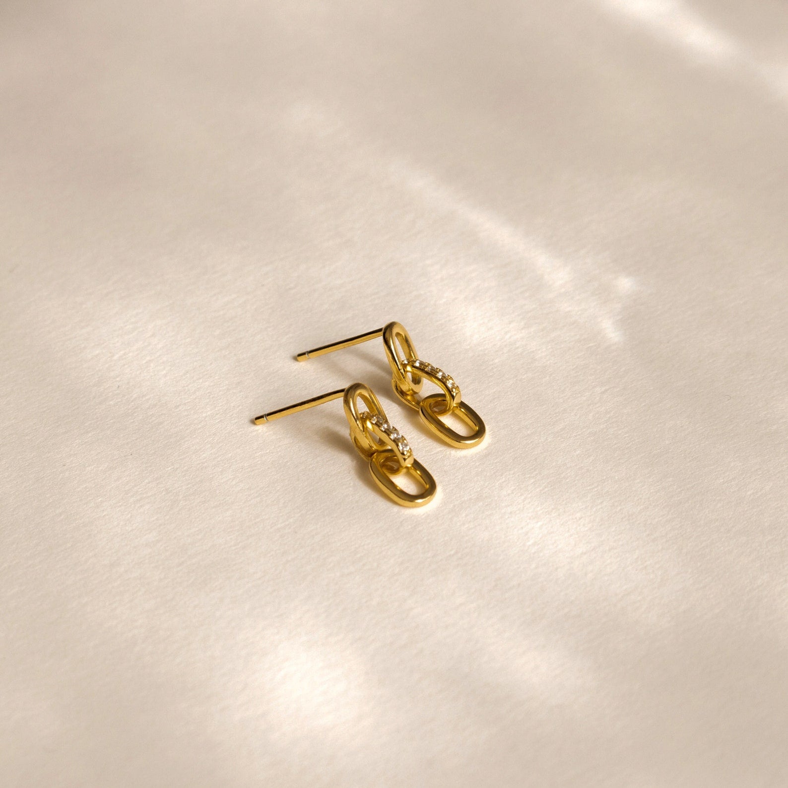 The Amia Pave Link Earrings, featuring small pavé accents, are displayed on a soft beige surface under gentle lighting.