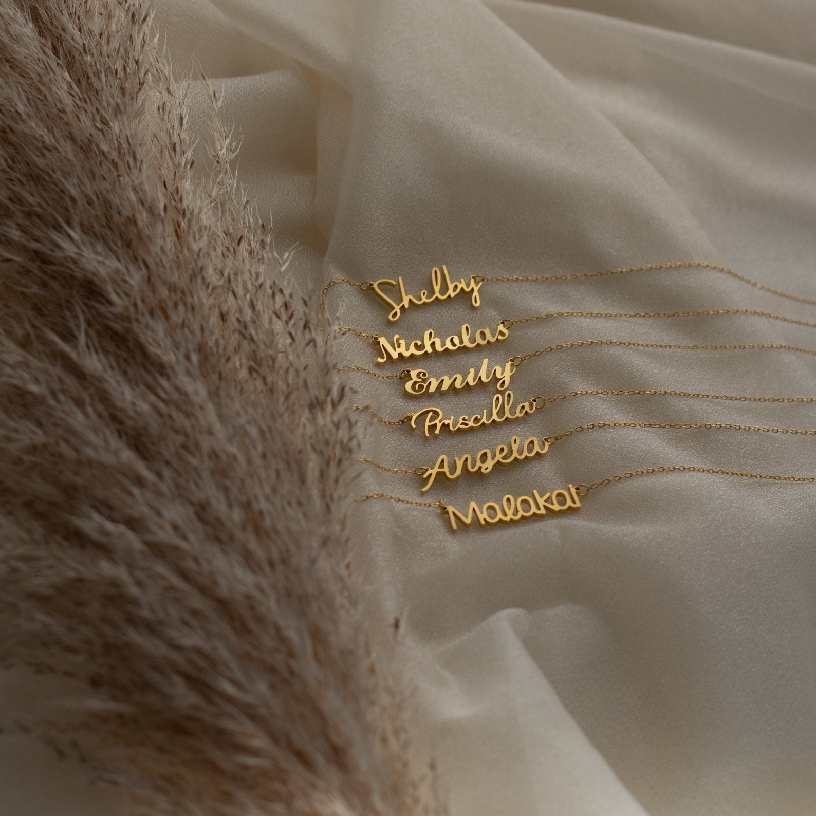 Five Nella Name Necklaces on cream fabric beside pampas grass, each custom nameplate displays Shelby, Nicholas, Emily, Priscilla, Angela, and Malaka—a stunning display of personalized jewelry.