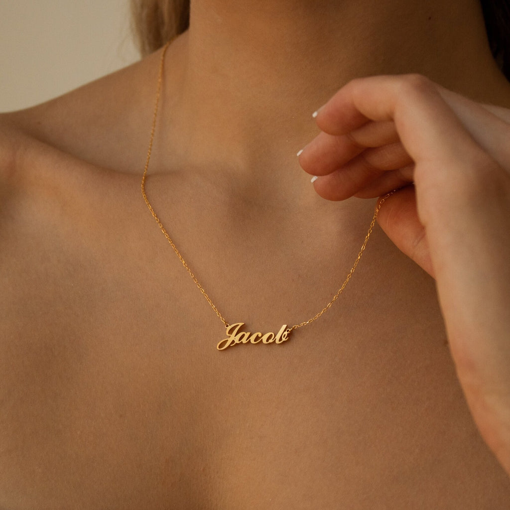 The Carrie Name Necklace in gold, featuring the name "Jacob" elegantly displayed and worn around a woman's neck, is a stunning example of personalized jewelry.