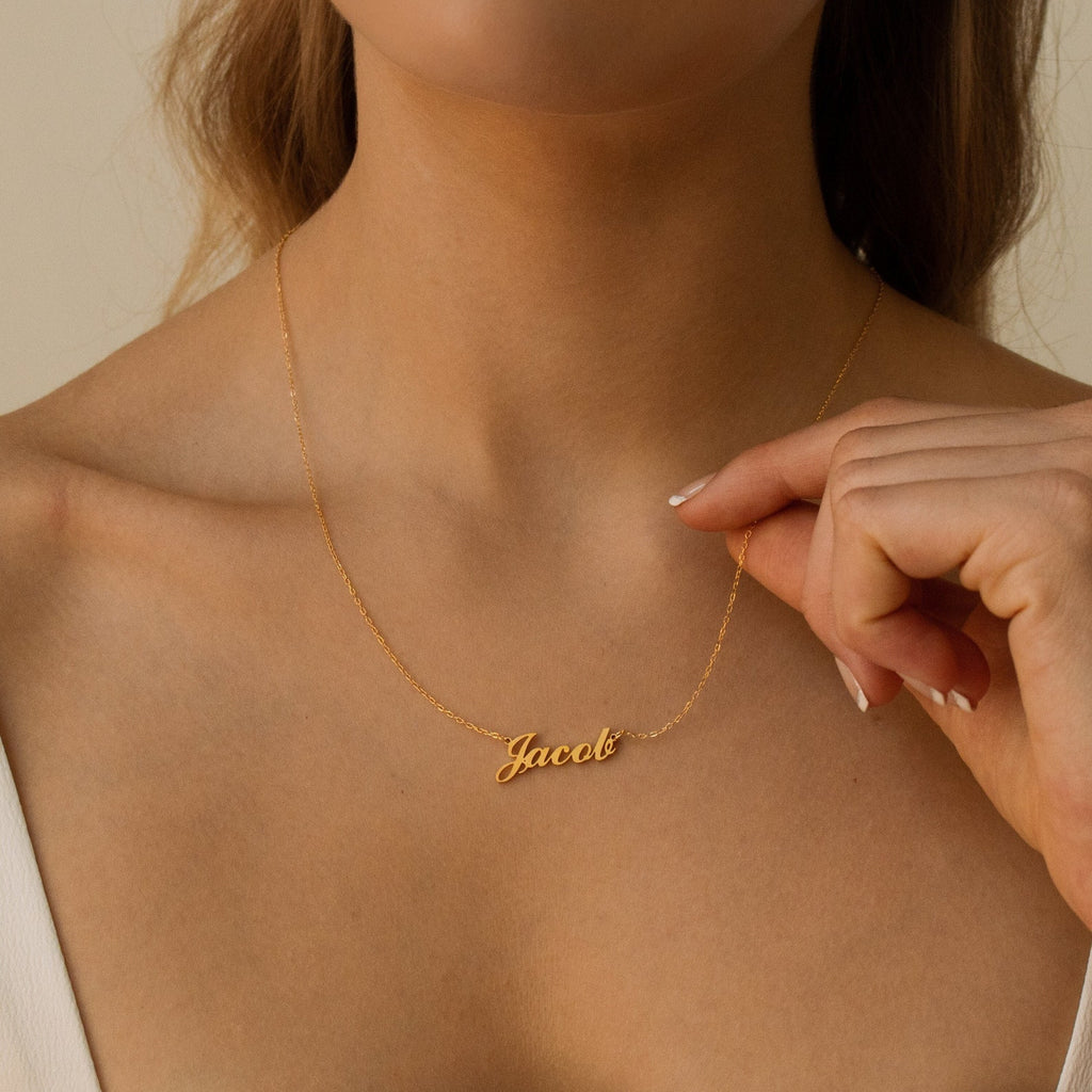 A woman holds a Carrie Name Necklace in gold, featuring the name "Jacob" in cursive, near her collarbone.