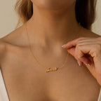 A woman holds a Carrie Name Necklace in gold, featuring the name "Jacob" in cursive, near her collarbone.