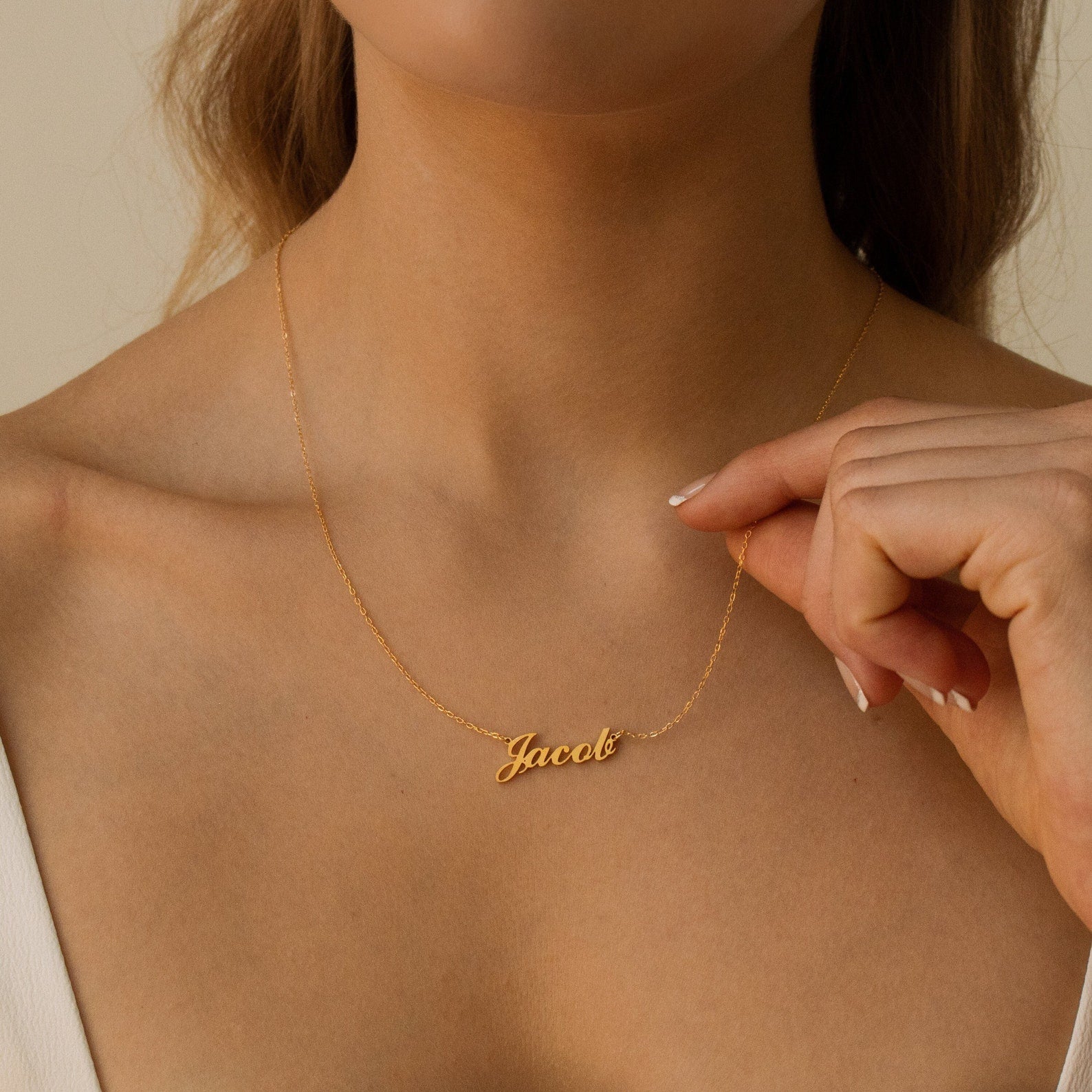 A woman holds a Carrie Name Necklace in gold, featuring the name "Jacob" in cursive, near her collarbone.