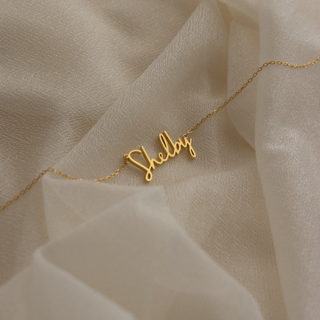 Nella Name Necklace featuring "Shelby" in elegant cursive, displayed on soft cream fabric—a beautiful personalized jewelry piece perfect for any occasion.