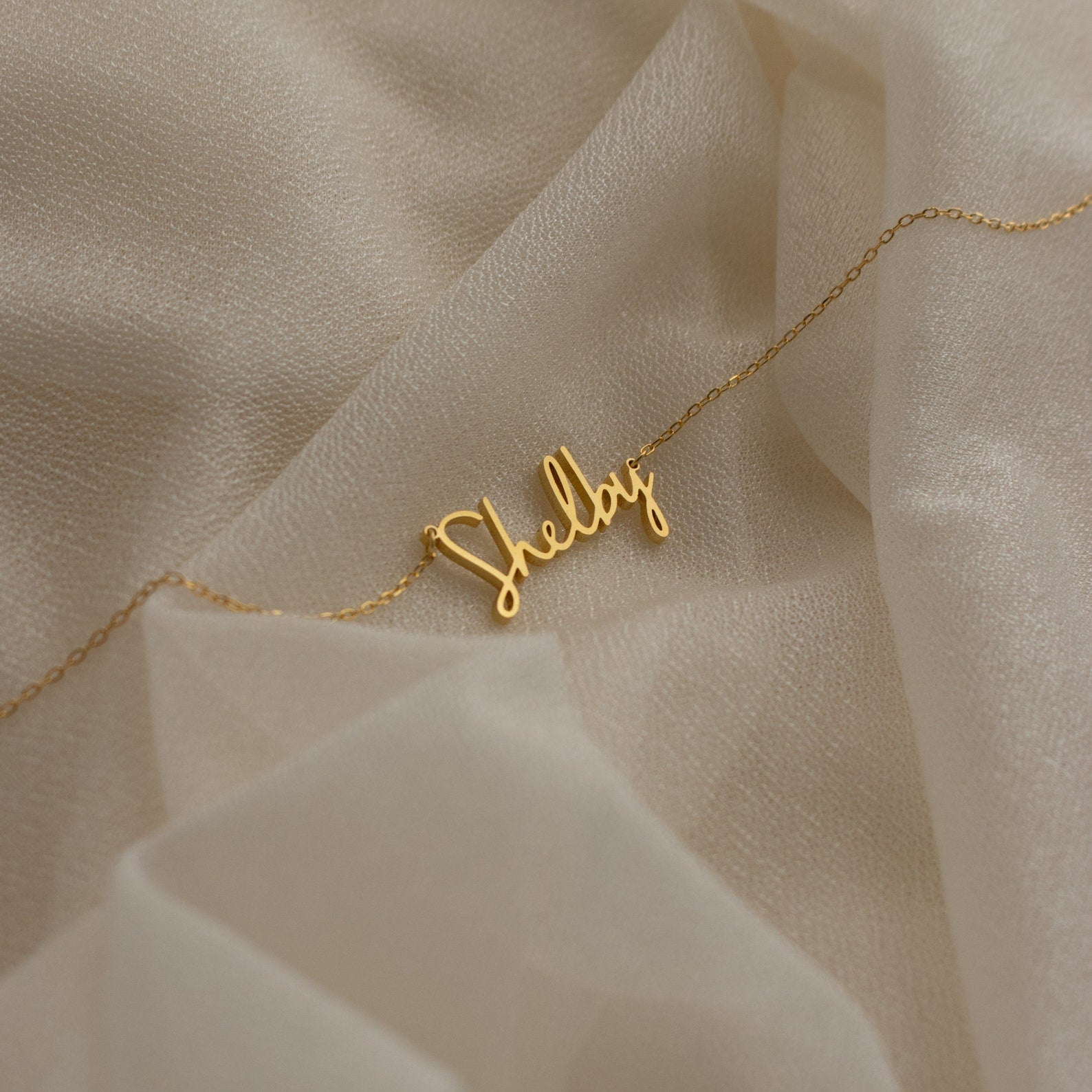 Nella Name Necklace featuring "Shelby" in elegant cursive, displayed on soft cream fabric—a beautiful personalized jewelry piece perfect for any occasion.