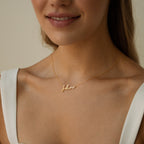 A woman wears a white top and the Nella Name Necklace, a gold custom nameplate featuring the name Jolene in elegant cursive script.