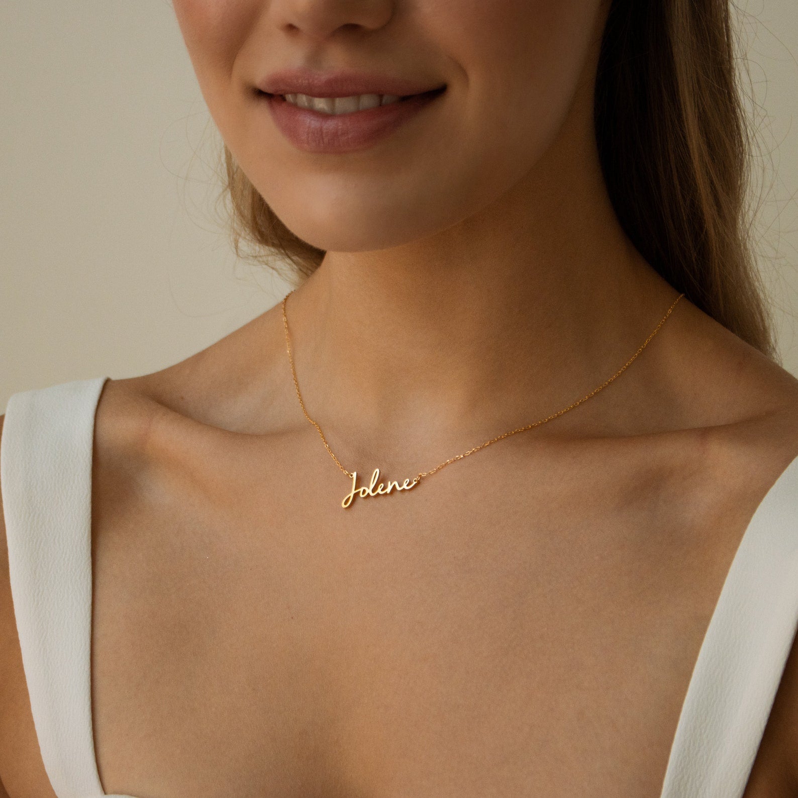 A woman wears a white top and the Nella Name Necklace, a gold custom nameplate featuring the name Jolene in elegant cursive script.