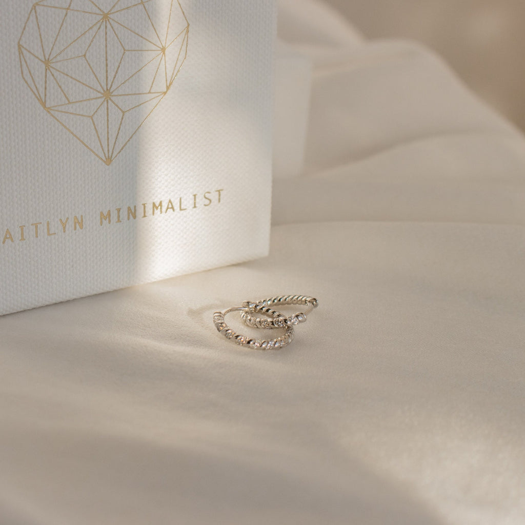 Two Aster Pave Twisted Hoops in silver sit on cream fabric next to a white box labeled AITLYN MINIMALIST—ideal for anyone who loves dainty earrings.
