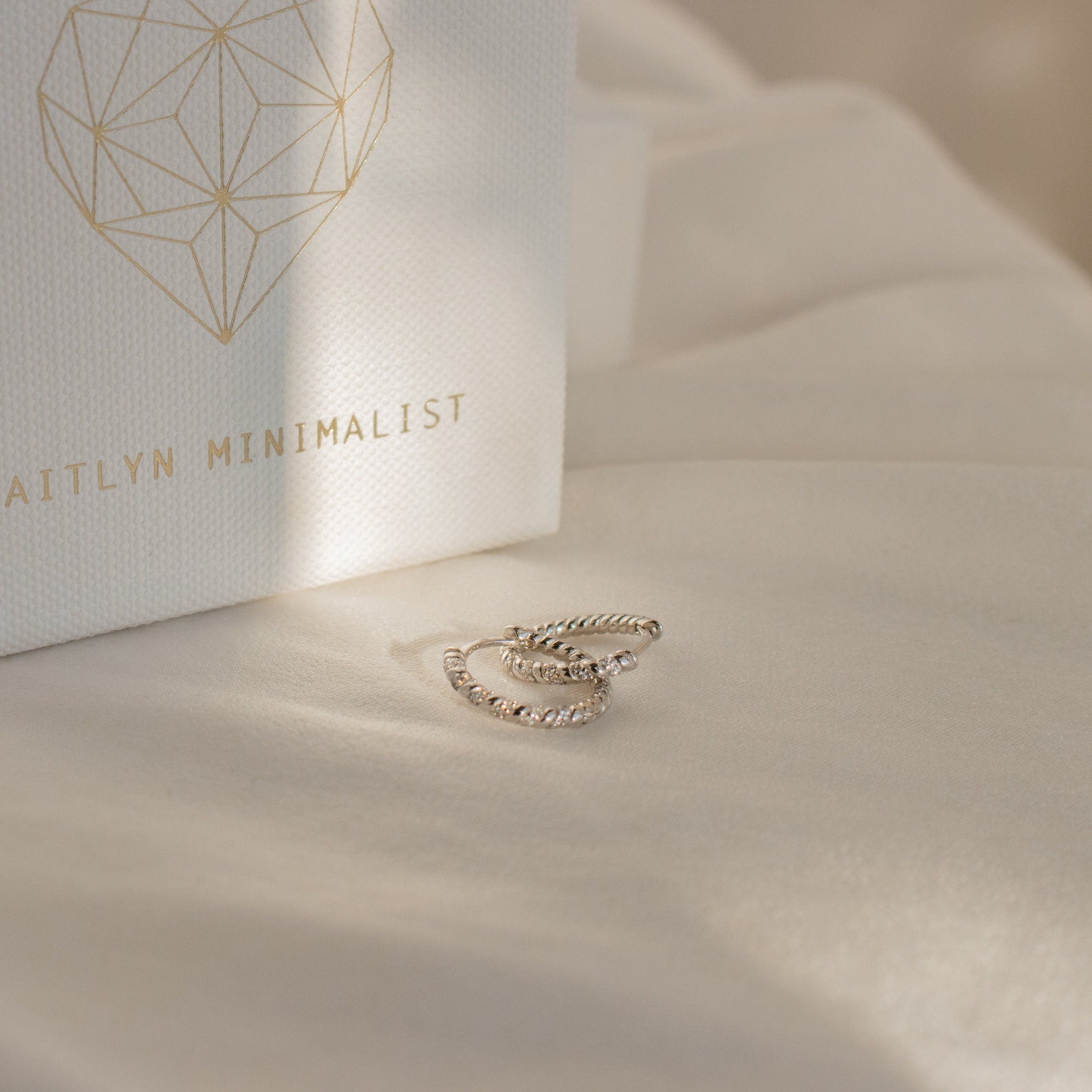 Two Aster Pave Twisted Hoops in silver sit on cream fabric next to a white box labeled AITLYN MINIMALIST—ideal for anyone who loves dainty earrings.