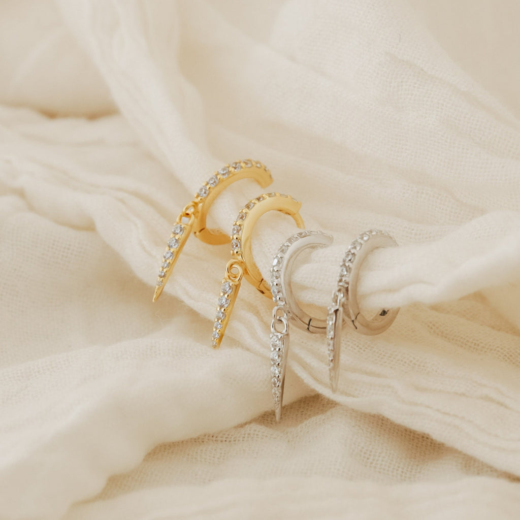 Two pairs of Quinn Pave Dagger Huggies—one gold, one silver—are displayed on soft cream-colored fabric, showcasing their edgy hoop designs that bring a bold accent to any outfit.