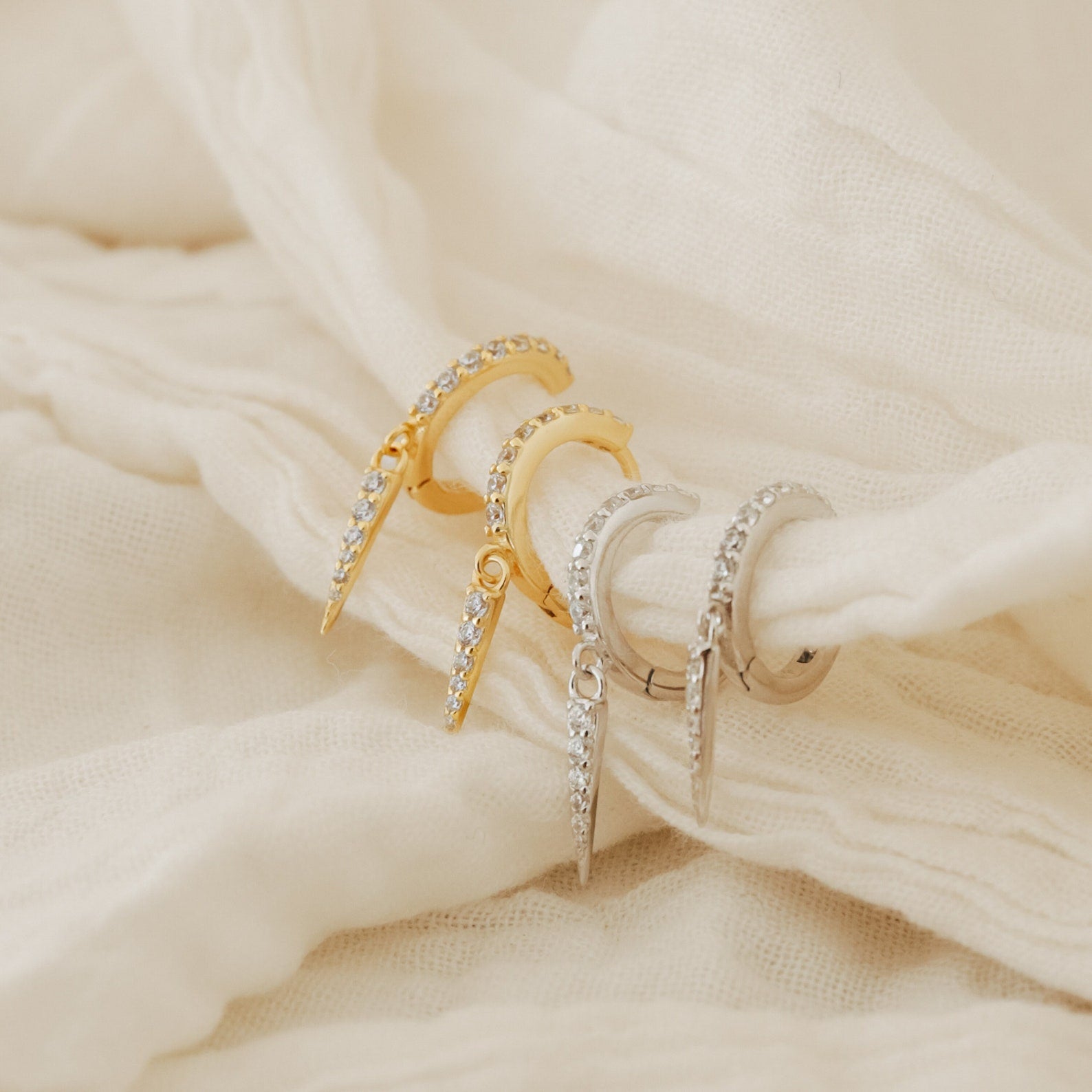 Two pairs of Quinn Pave Dagger Huggies—one gold, one silver—are displayed on soft cream-colored fabric, showcasing their edgy hoop designs that bring a bold accent to any outfit.