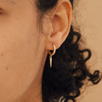 A close-up of a person wearing the Quinn Pave Dagger Huggies, which feature gold hoops with small spike pendants on their ear.