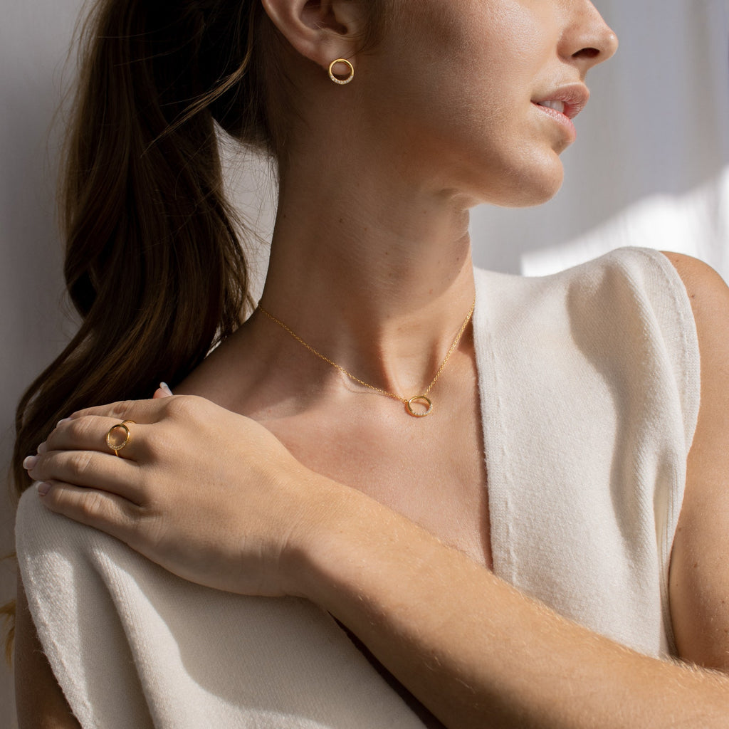 A woman in a sleeveless cream top looks to the side, wearing minimalistic gold circle earrings, a ring, and the Kayla Pave Circle Necklace.
