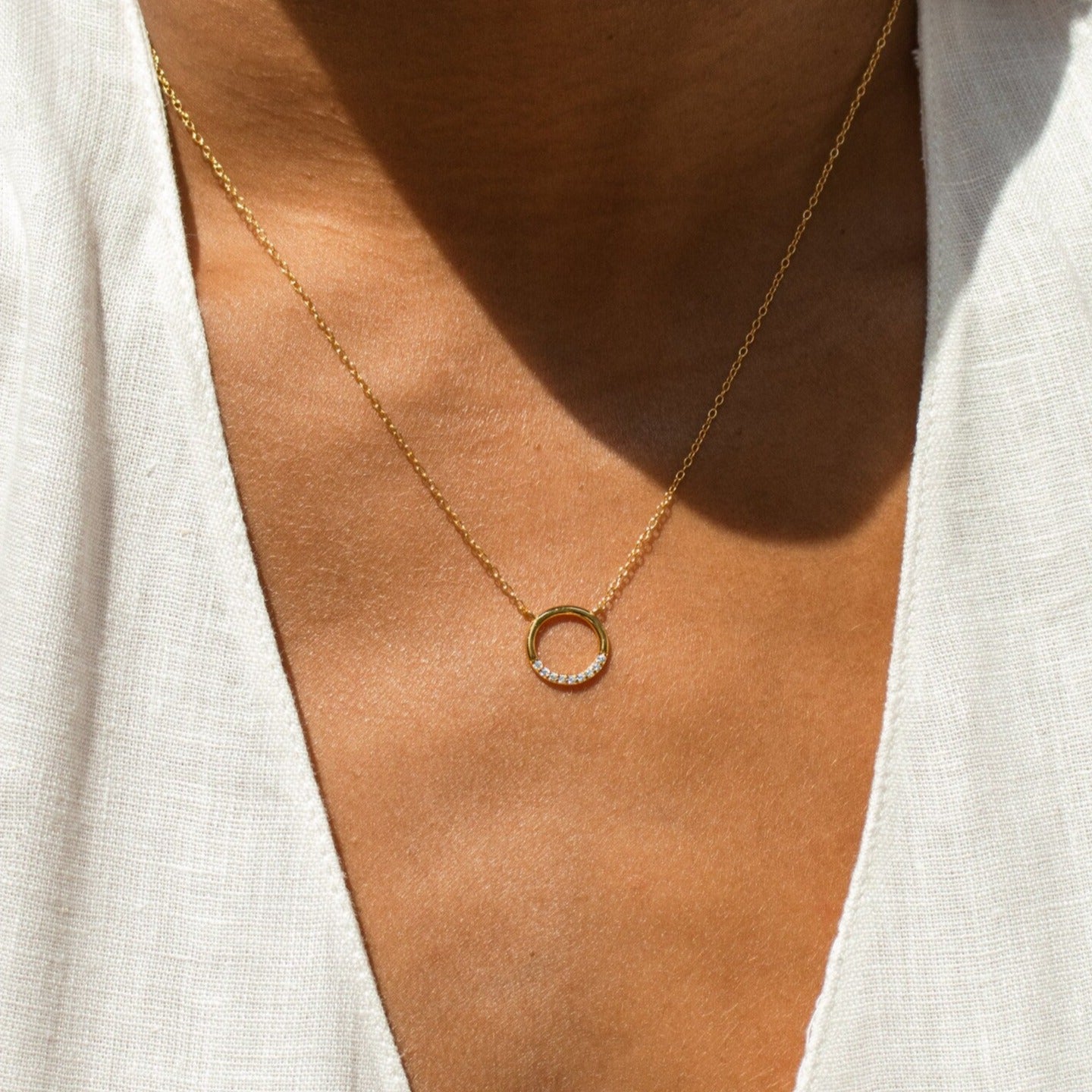 Minimal gold circle necklace featuring a row of small white stones along the lower arc, worn by a model in a white V-neck top.
