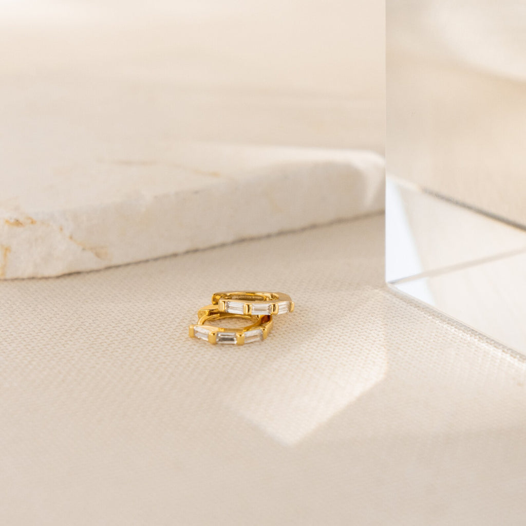 The Kenia Diamond Huggies, two small gold hoop earrings with rectangular gems inspired by classic baguette diamond earrings, rest on a light textured surface beside a marble and glass block.