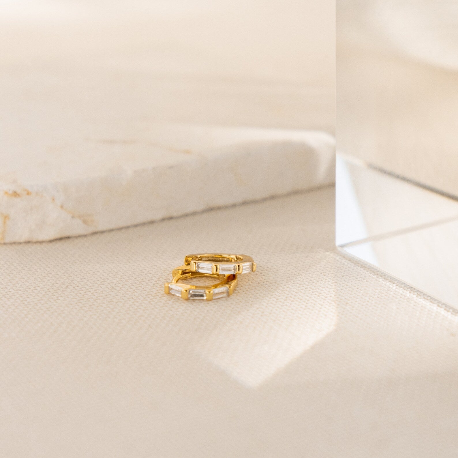 The Kenia Diamond Huggies, two small gold hoop earrings with rectangular gems inspired by classic baguette diamond earrings, rest on a light textured surface beside a marble and glass block.