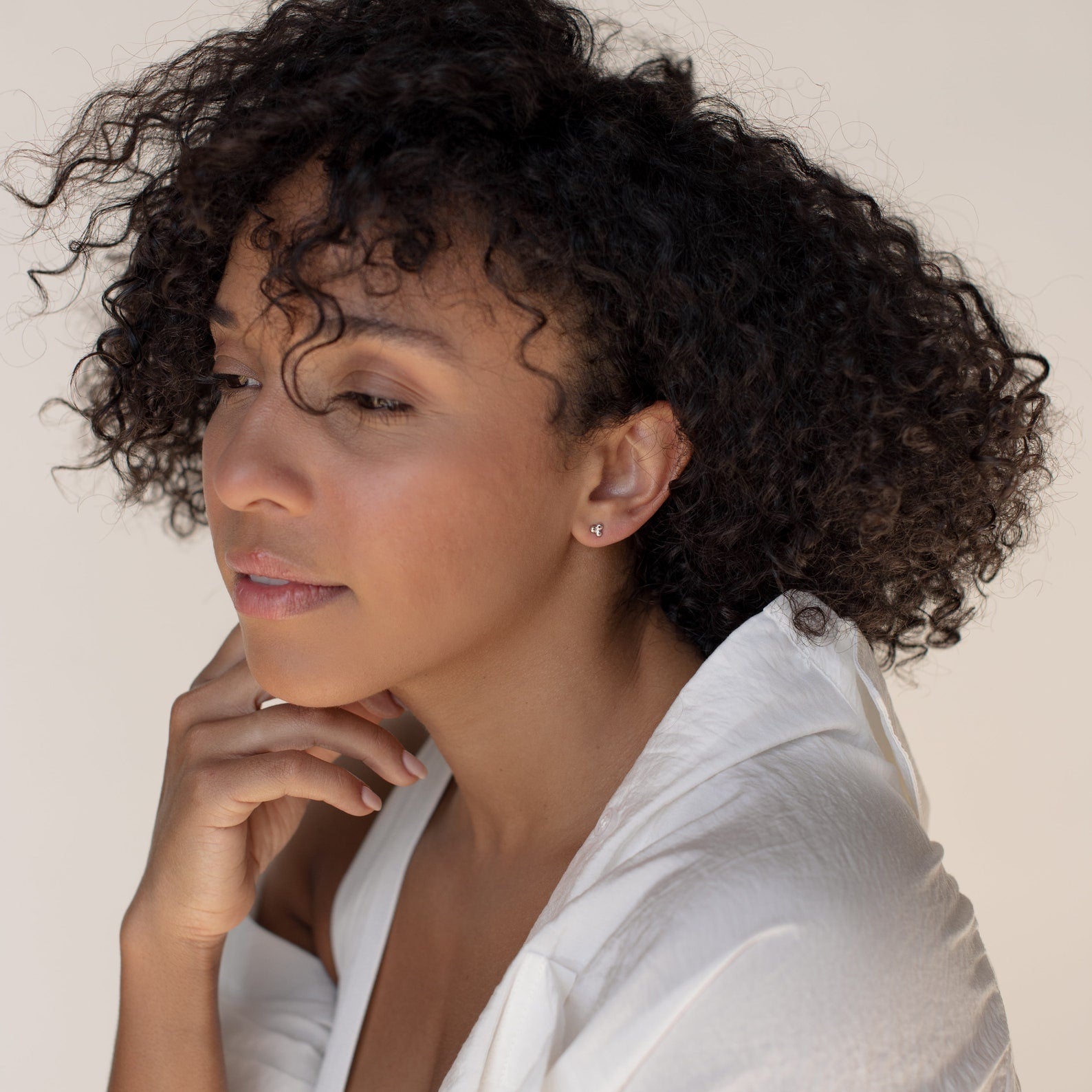A woman with curly hair in a white shirt looks thoughtful, resting her chin on her hand and showcasing an elegant earring stack featuring the Trio Sphere Studs.