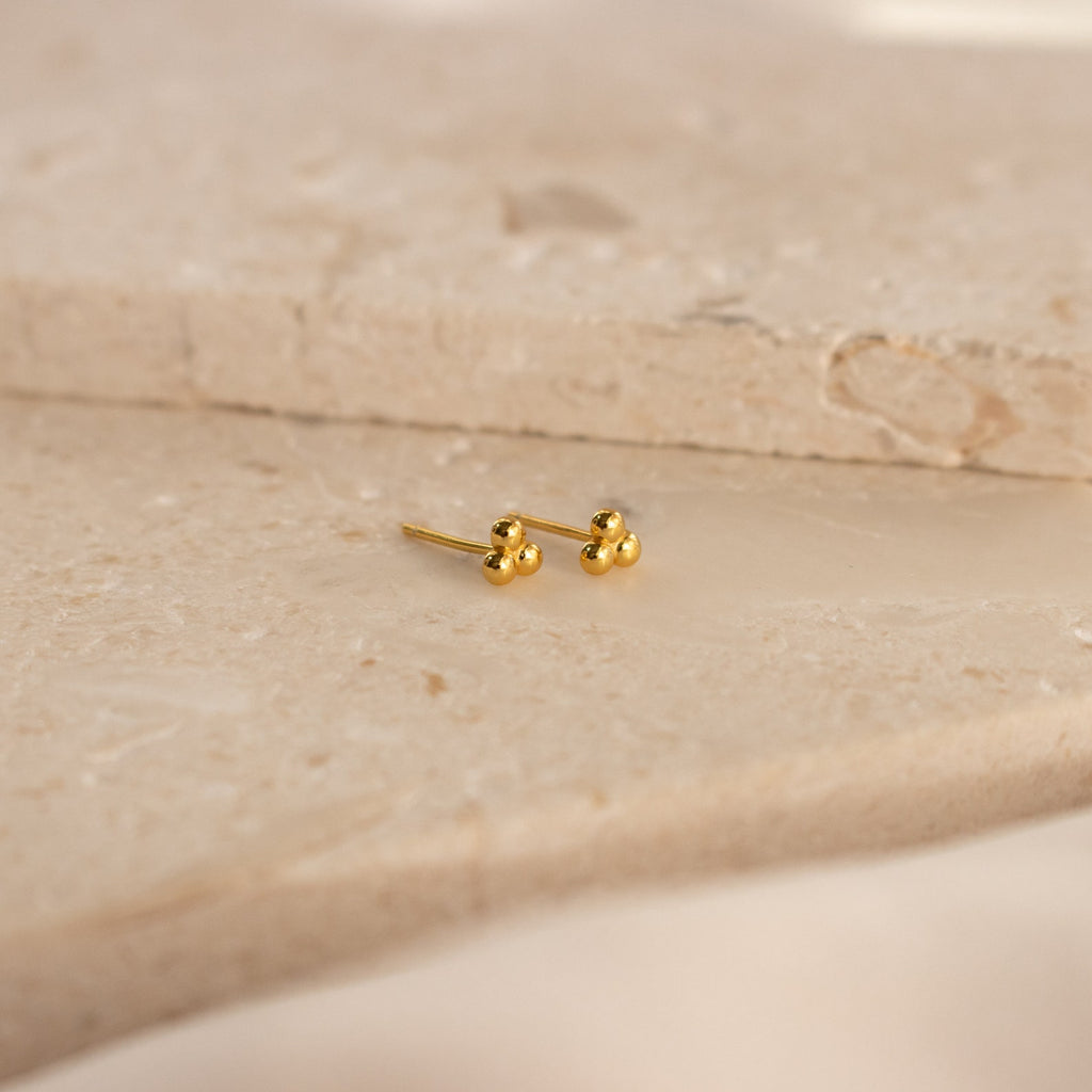 The Trio Sphere Studs feature a small gold three-dot cluster design, displayed on a beige stone surface—ideal for creating an elegant earring stack or accenting alternative piercings.