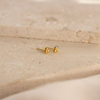 The Trio Sphere Studs feature a small gold three-dot cluster design, displayed on a beige stone surface—ideal for creating an elegant earring stack or accenting alternative piercings.