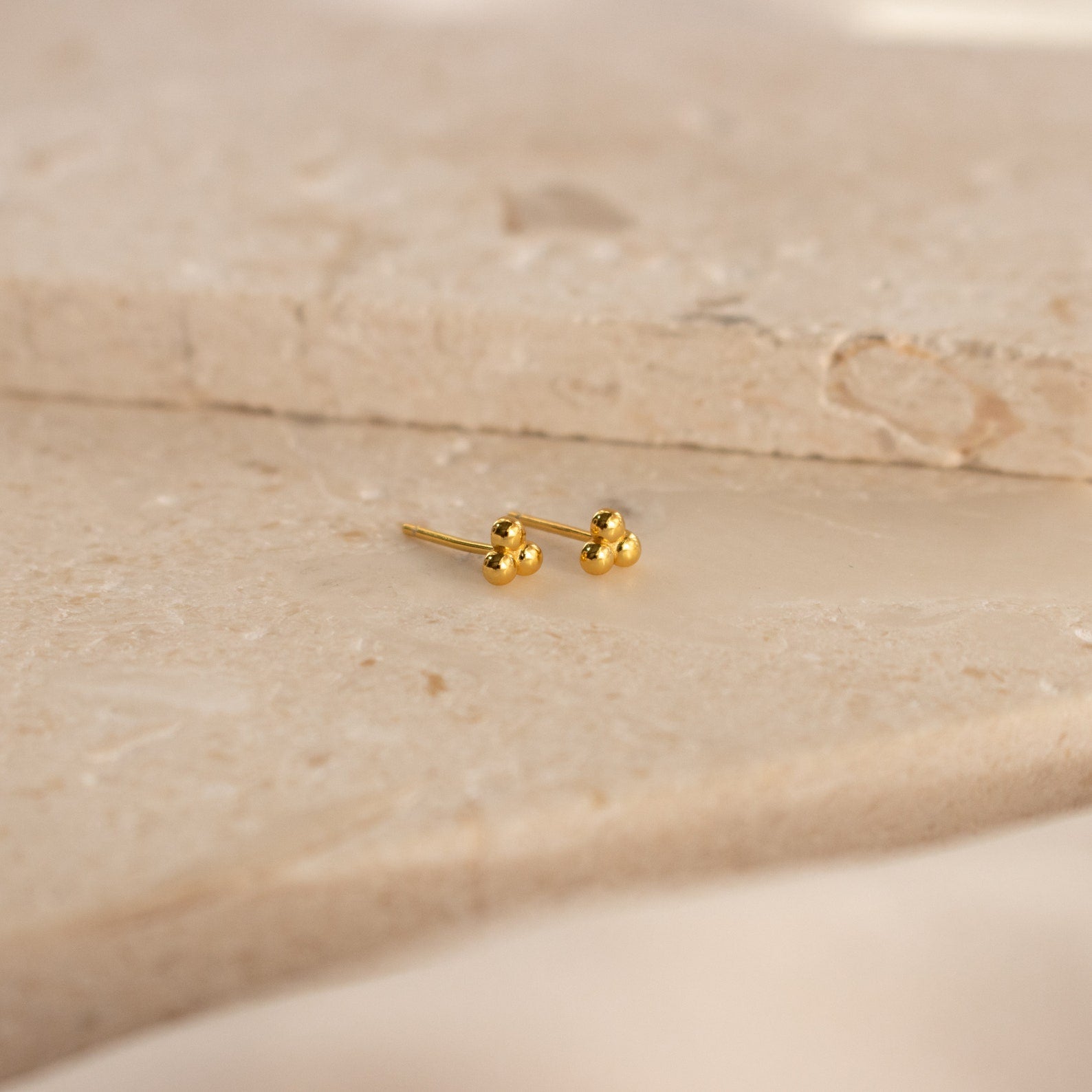 The Trio Sphere Studs feature a small gold three-dot cluster design, displayed on a beige stone surface—ideal for creating an elegant earring stack or accenting alternative piercings.