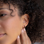 Close-up of a person with curly hair touching the Black Enamel Huggies earring on their ear.