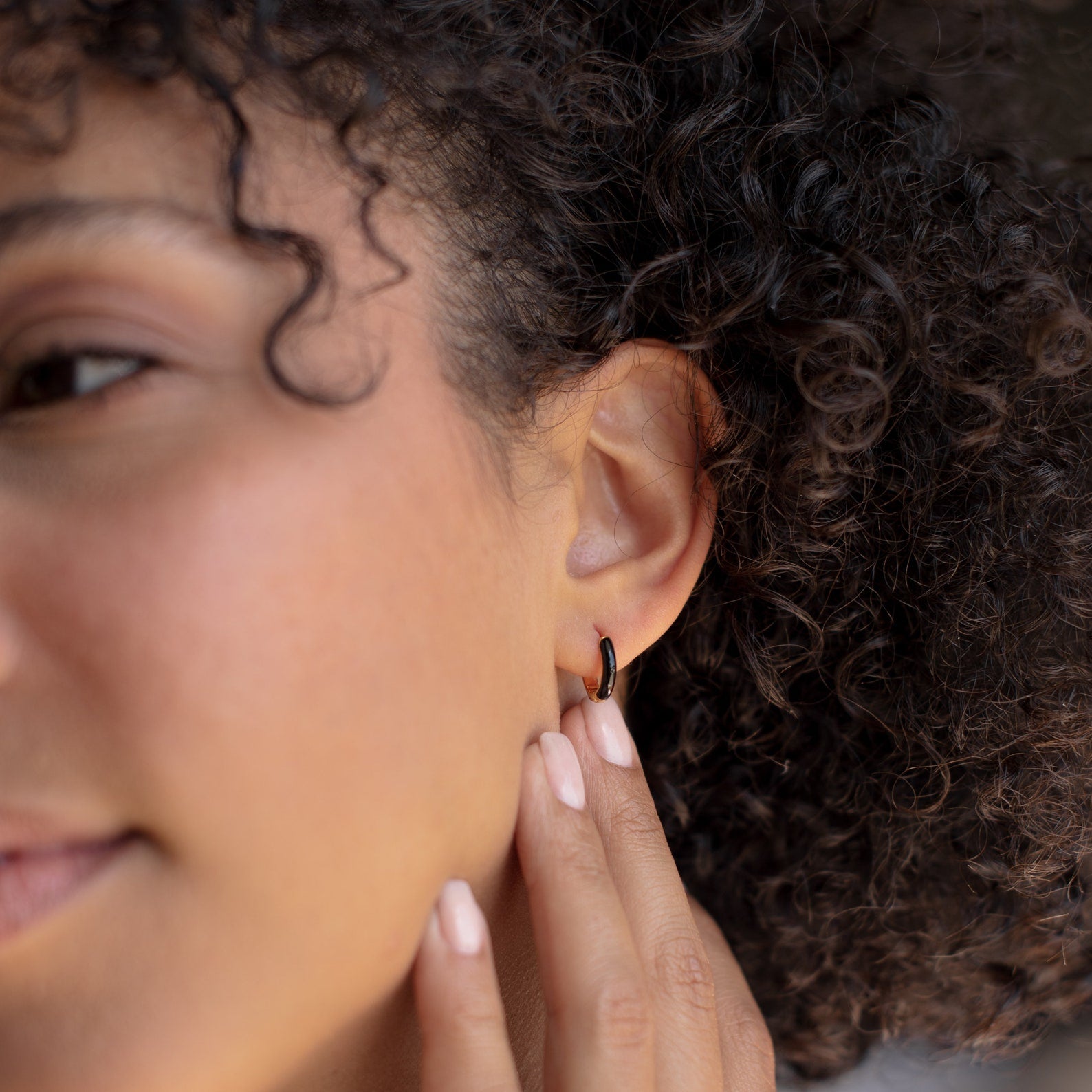 Close-up of a person with curly hair touching the Black Enamel Huggies earring on their ear.