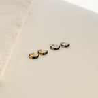 Four small hoop earrings—two gold, two silver—featuring the Black Enamel Huggies are displayed on a minimalist, light surface.