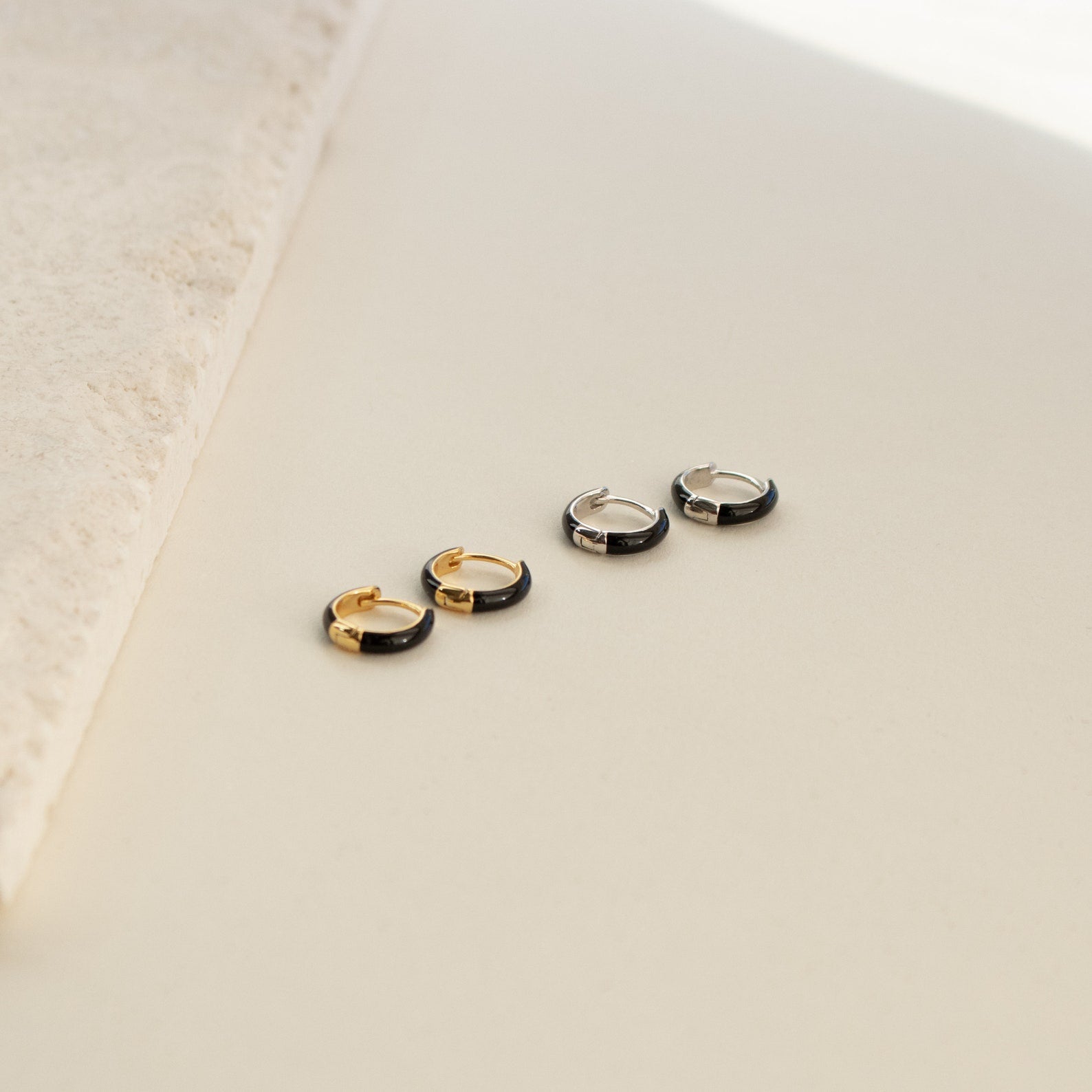 Four small hoop earrings—two gold, two silver—featuring the Black Enamel Huggies are displayed on a minimalist, light surface.