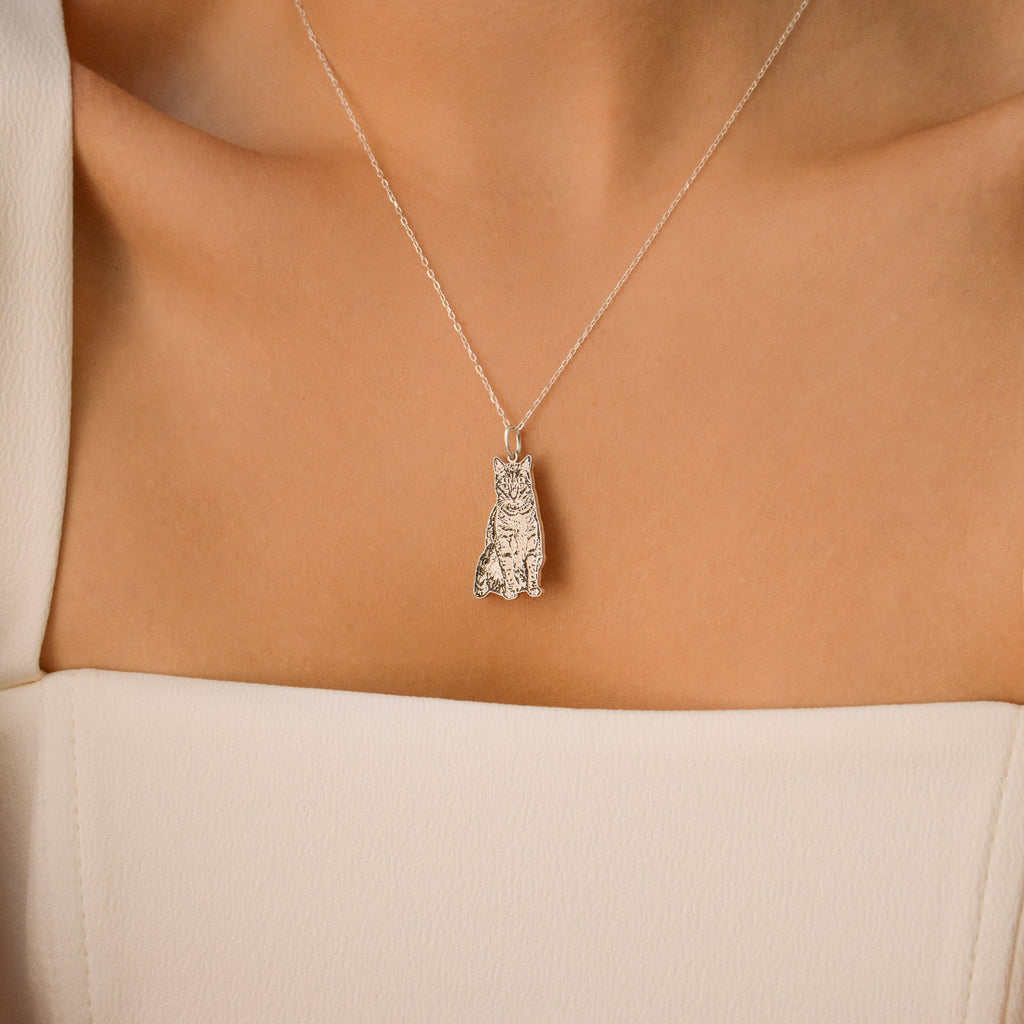 A Custom Pet Portrait Necklace featuring a silver cat pendant amulet is worn on a woman's neck, paired with a white off-shoulder top.