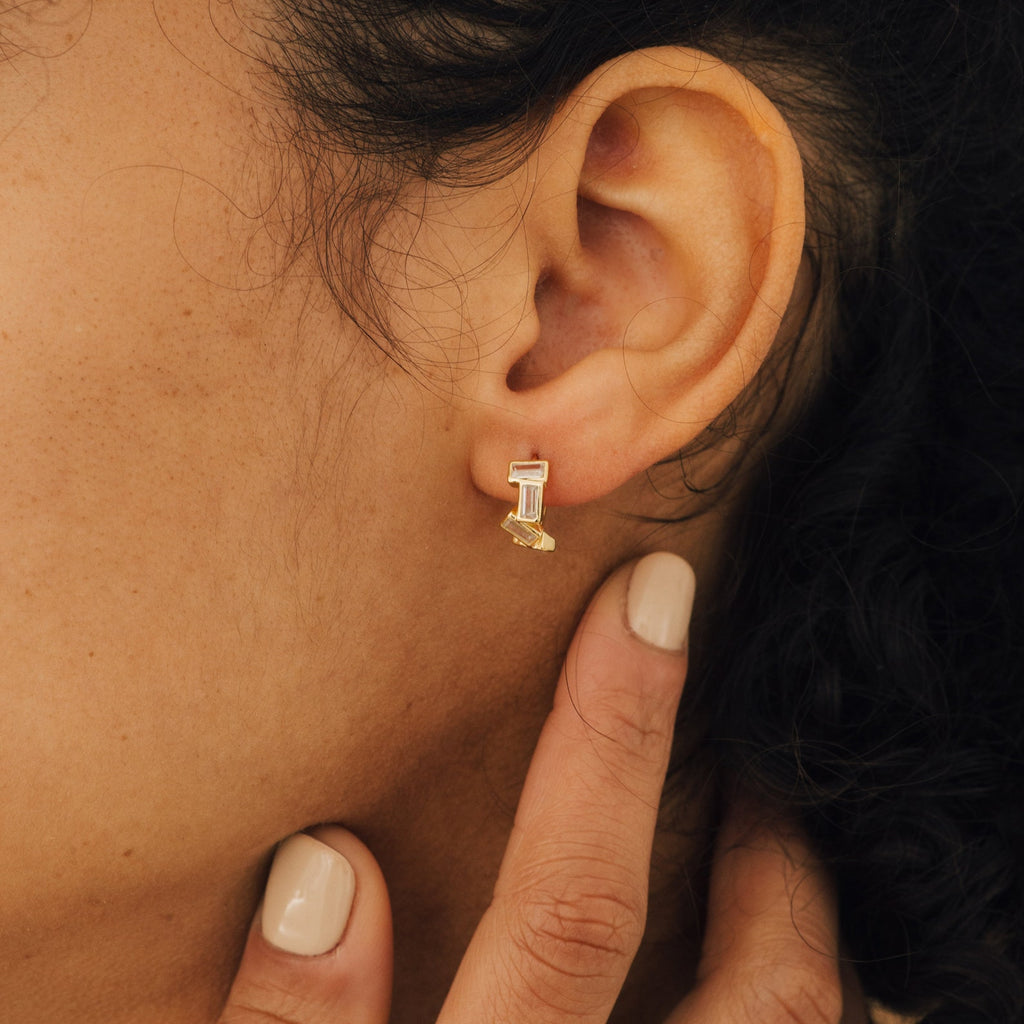 A close-up shows someone wearing Glacier Baguette Huggies, their fingers gently touching their neck to highlight the minimalistic sparkle and elegant gold geometric design of these earrings.