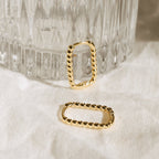 The Indiana Beaded Hoops, stylish gold rectangle earrings with elegant beaded details, are displayed on a white cloth beside a textured glass for a chic look.