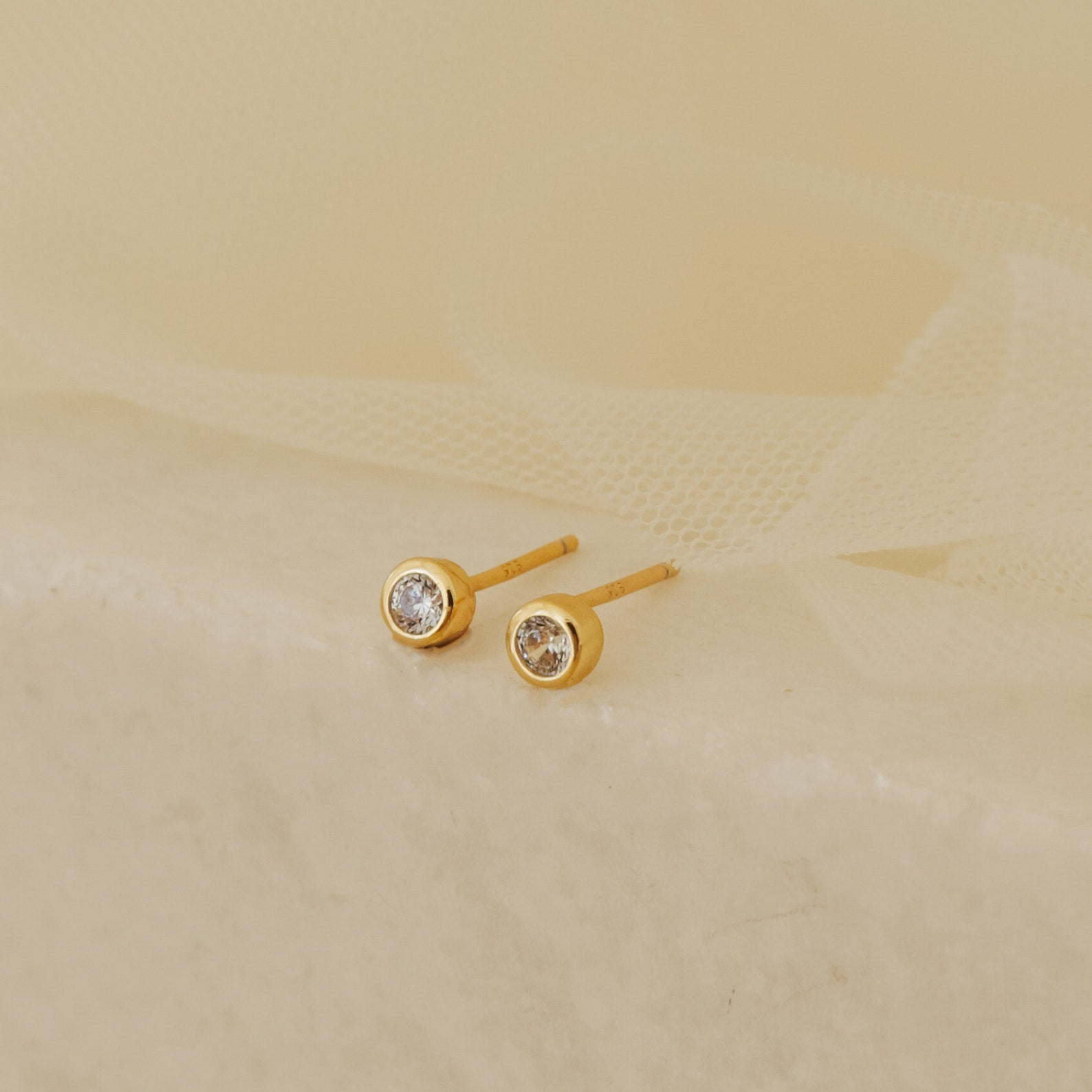 Jane Diamond Studs, featuring small round brilliant diamonds, are elegantly displayed on a soft beige fabric background.