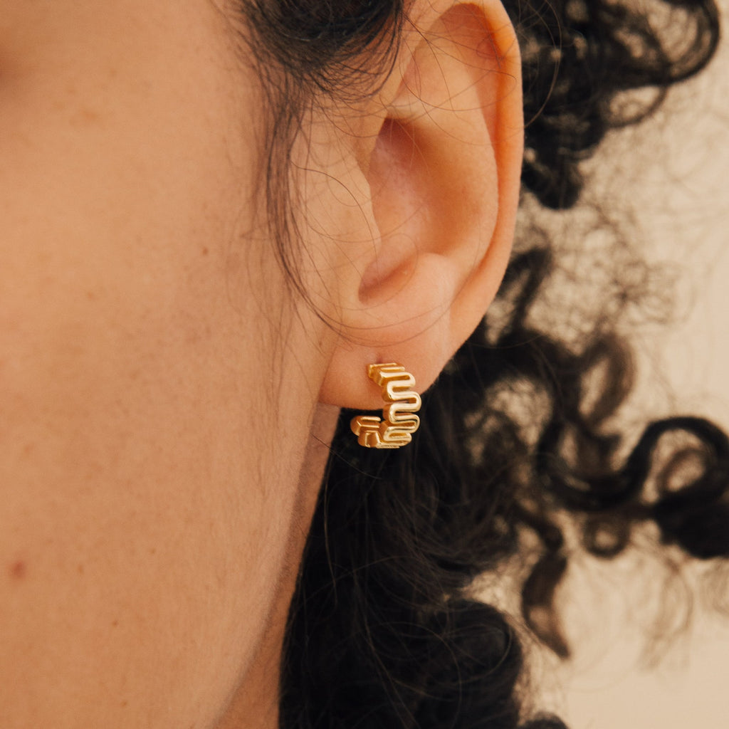 Close-up of an ear with curly hair, wearing gold Maze Half Hoops—wavy-shaped earrings ideal for creating a unique earring stack.