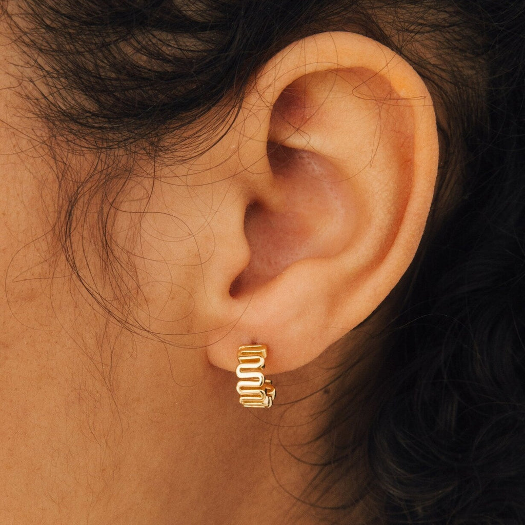 Close-up of an ear wearing a small, gold, wavy-patterned hoop earring—Maze Half Hoops are perfect for adding standout style to your unique earring stack.