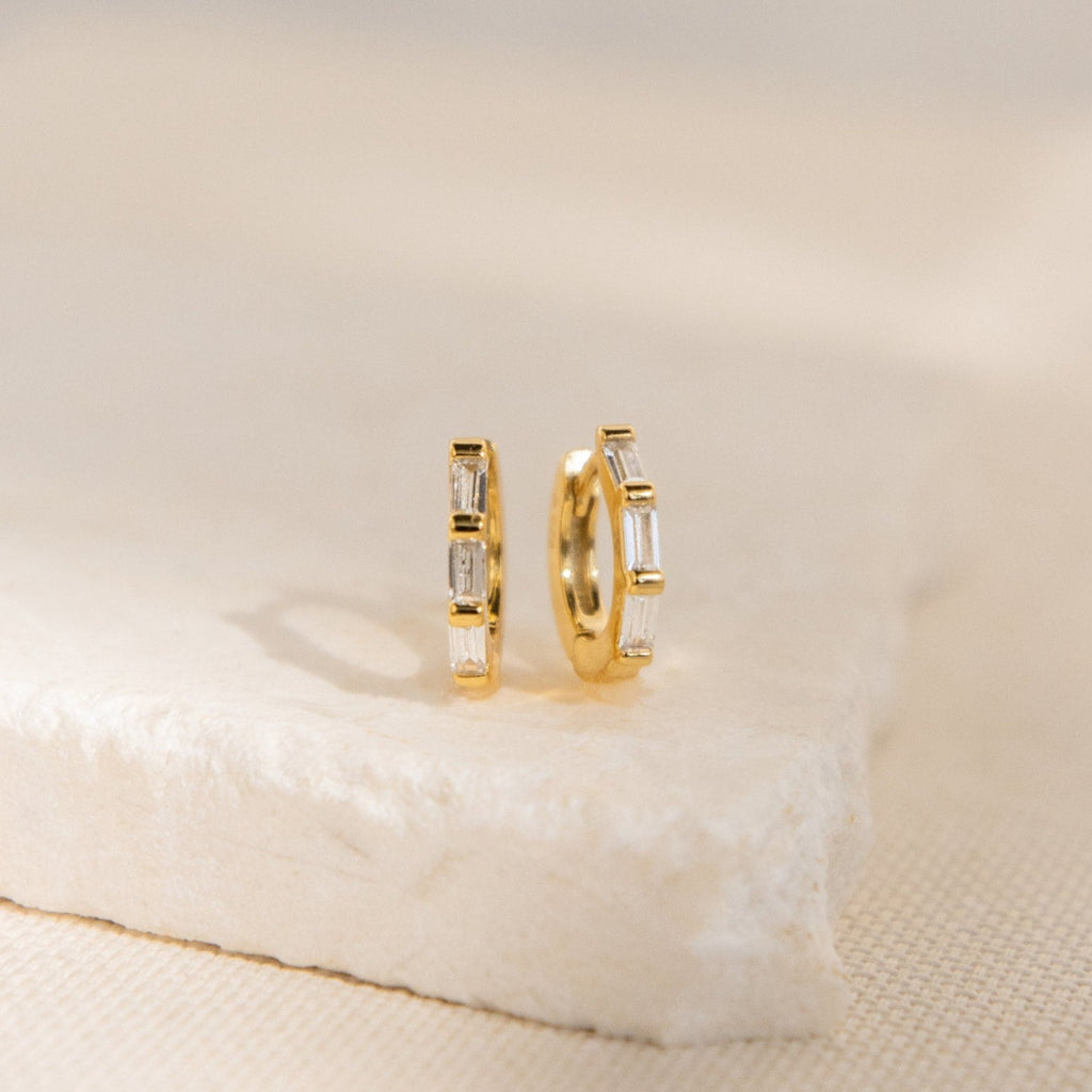 The Kenia Diamond Huggies are small gold earrings with rectangular clear stones, shown upright on a light textured surface—ideal as elegant wedding earrings or chic baguette diamond accessories.